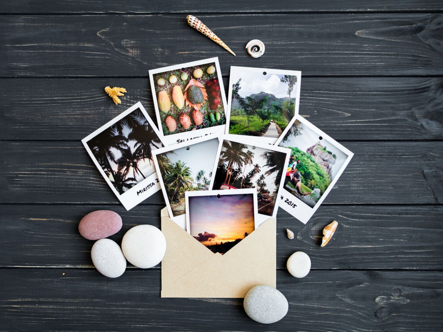 Polaroid photos spilling from an envelope on a dark wooden surface, surrounded by shells and stones.
