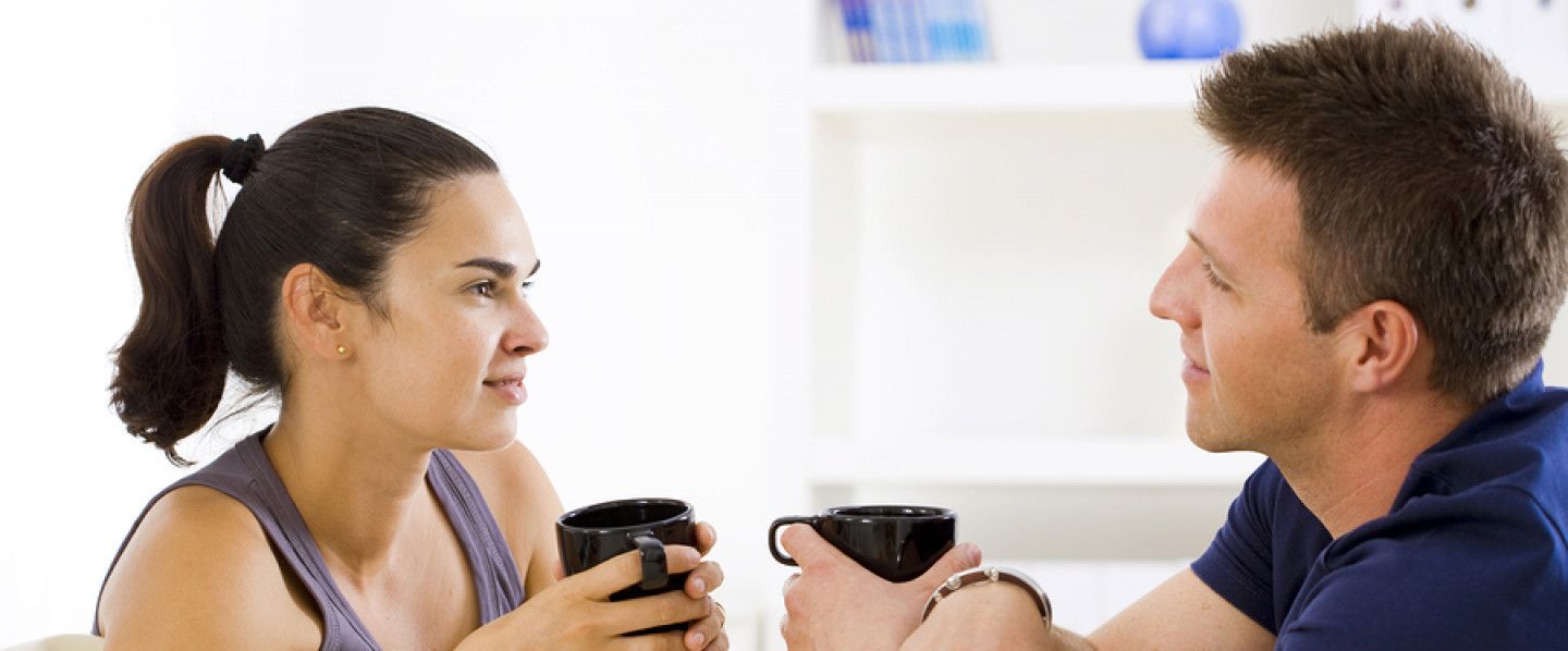 A couple, side-by-side, drinks coffee and looks at each other. They are indoors.