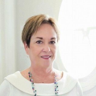 Woman with short brown hair, smiling, wearing a white top and beaded necklace.