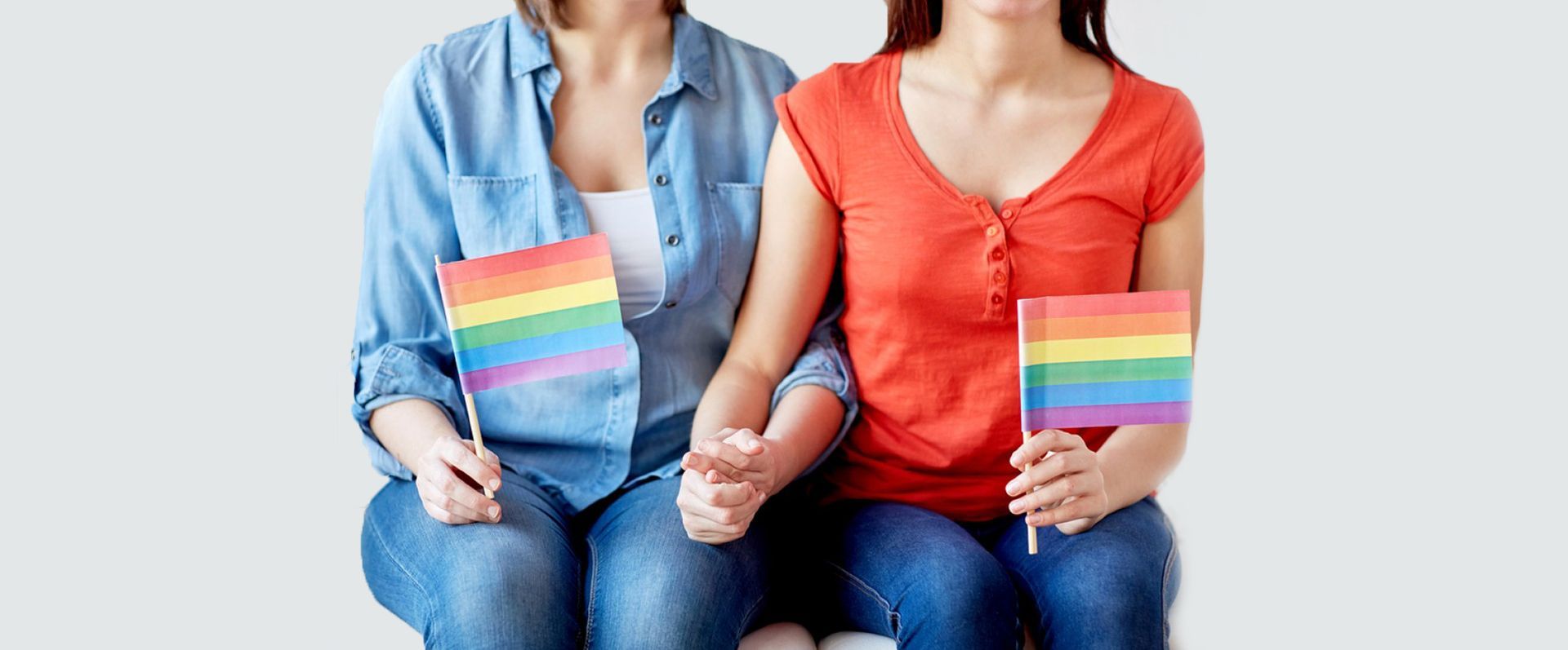 Two women holding rainbow flags, sitting close and holding hands, in jeans and t-shirts.