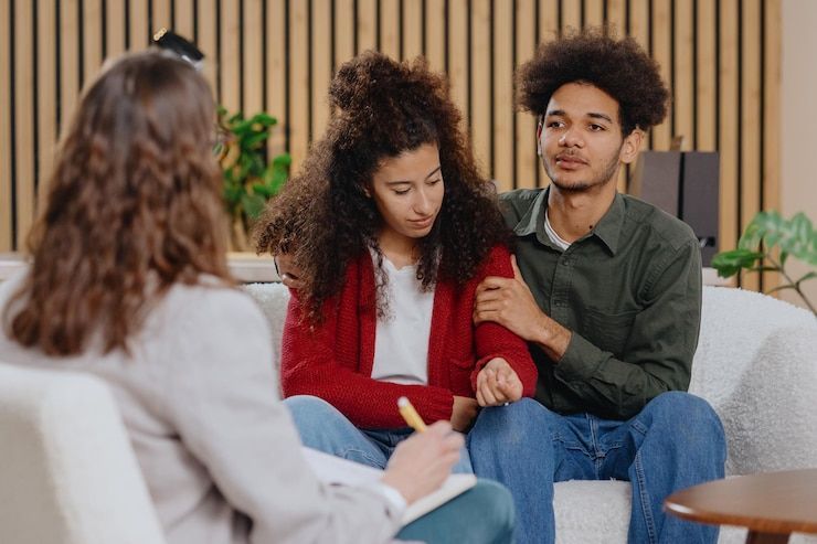 Couple holding hands during therapy session, seated on a couch; therapist's hand holding a clipboard.