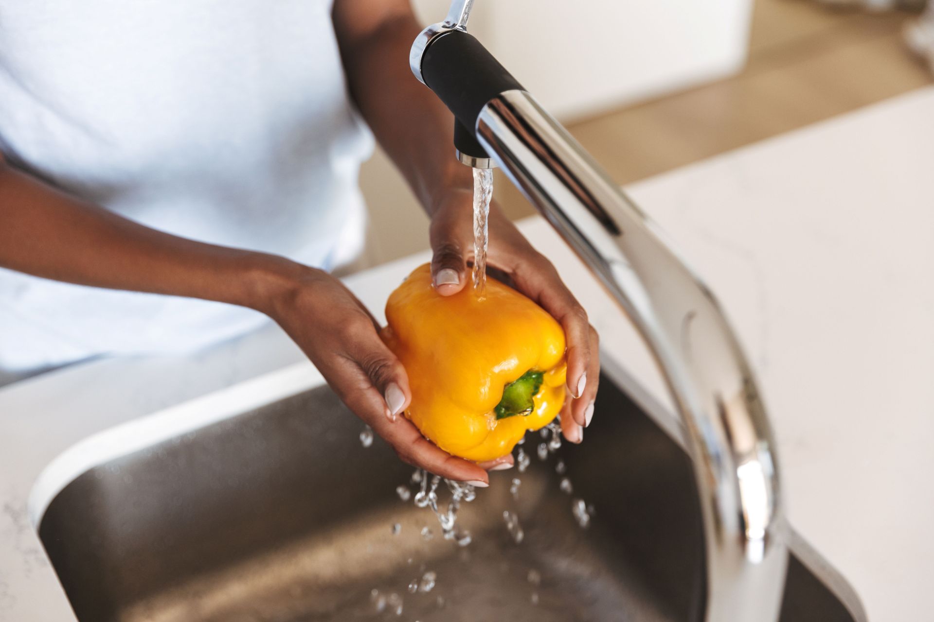 Hands rinsing a yellow bell pepper under a kitchen faucet at a stainless steel sink.