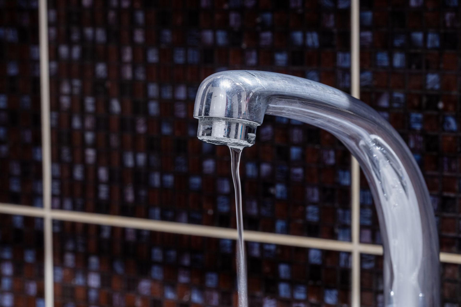 Close-up of a faucet with a slow, thin stream of water against a dark tiled backsplash.