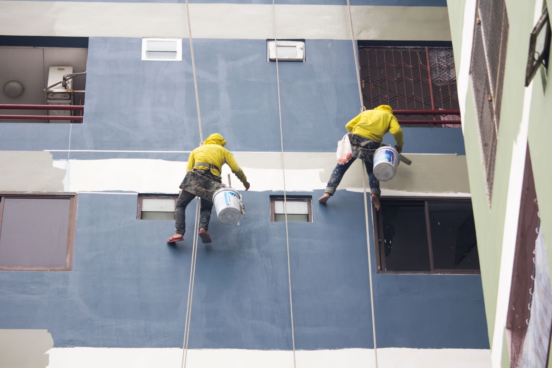 Two painters in yellow suits hanging on ropes, painting a tall building.