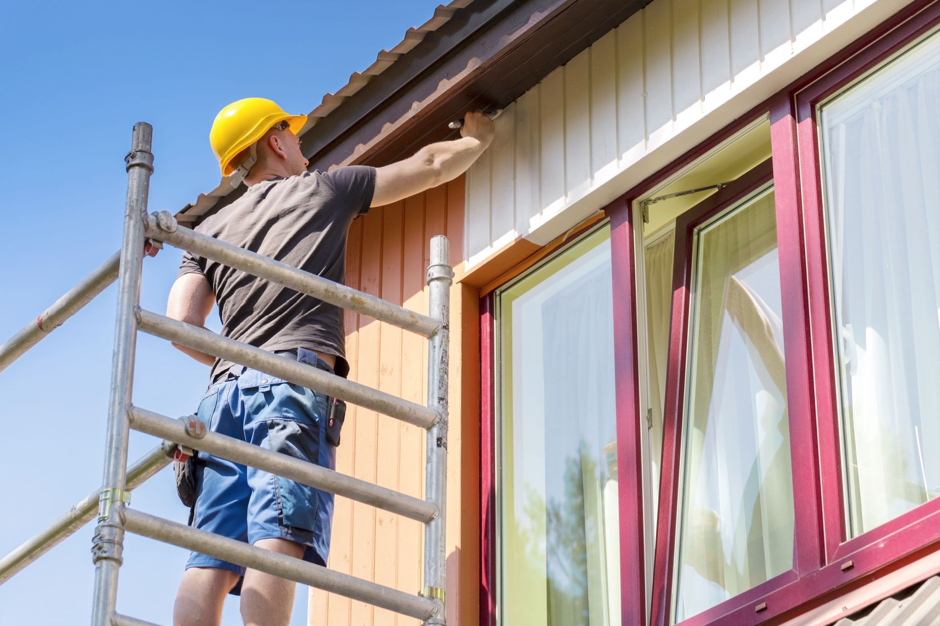 Person painting house trim while standing on a ladder under a blue sky.