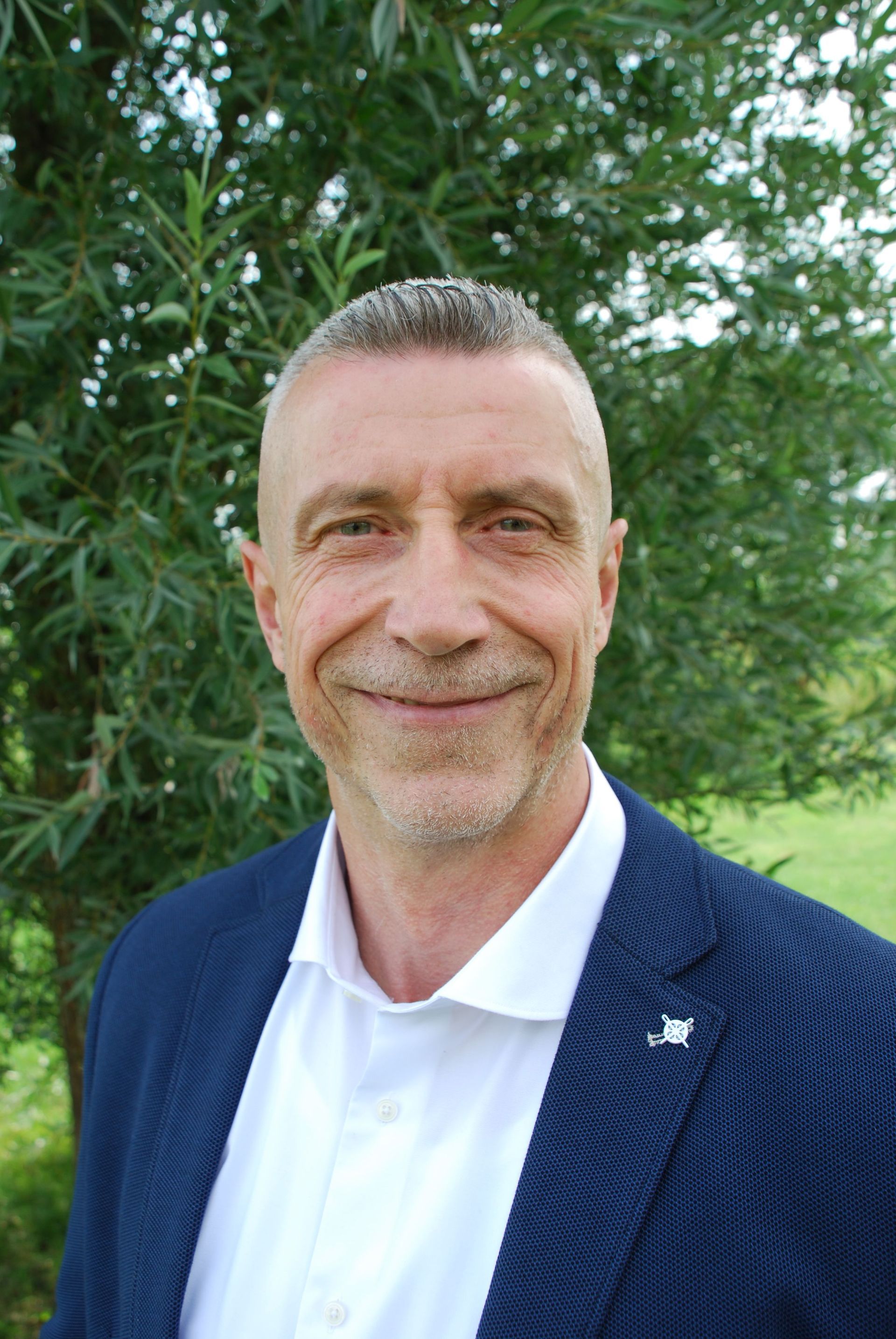 Een lachende man in een donkerblauwe blazer en witte polo staat buiten voor groene bomen.