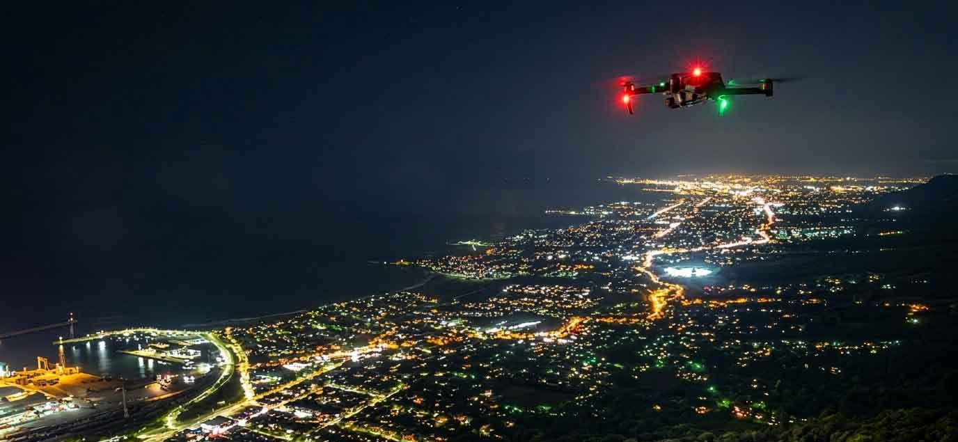 Drone with red and green lights flying over a city at night, with illuminated streets and coastline below