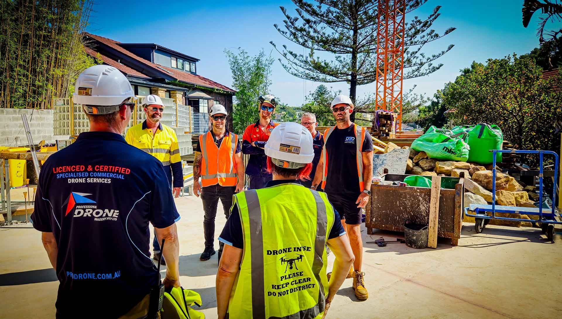 Construction crew in safety vests and hard hats gathered on a site beside equipment and building materials.