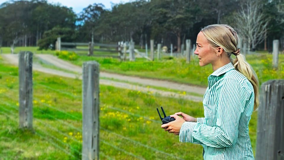 Port Macquarie NSW Farmers Training Person in a green striped shirt holding a drone controller beside a fenced field with grazing horses.