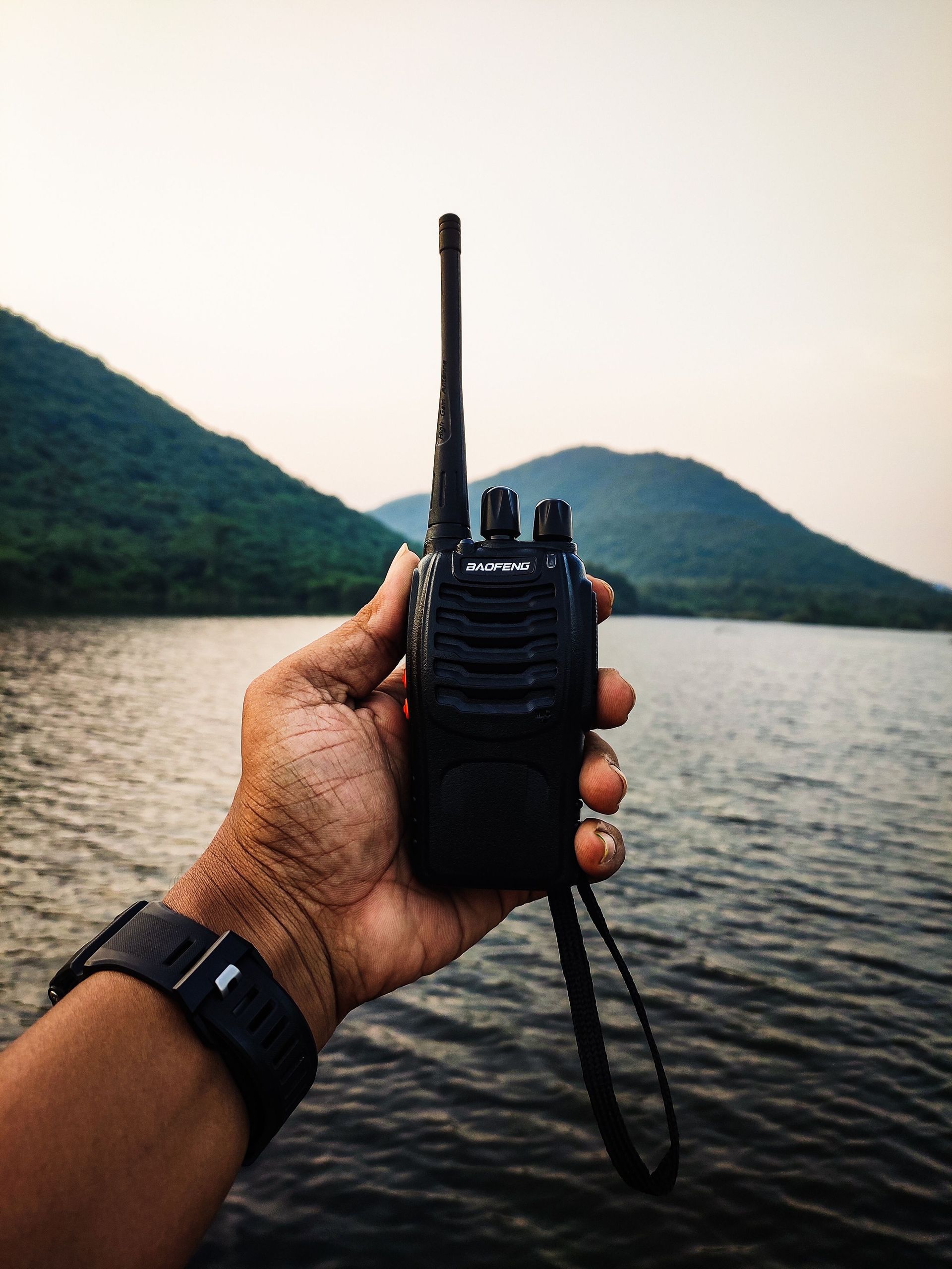 Hand holding a black walkie-talkie by a calm lake with green hills in the background