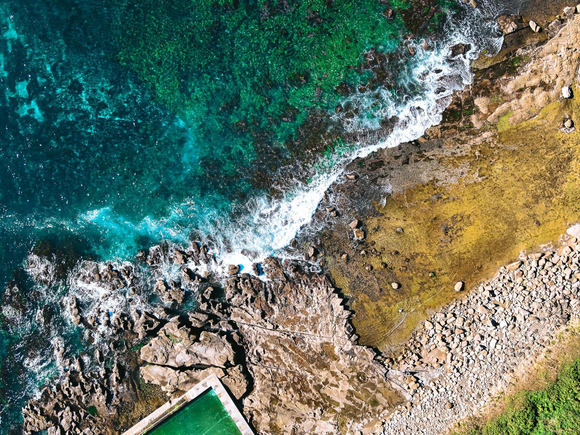 Aerial view of turquoise waves crashing against a rocky shoreline with patches of yellow-brown land.