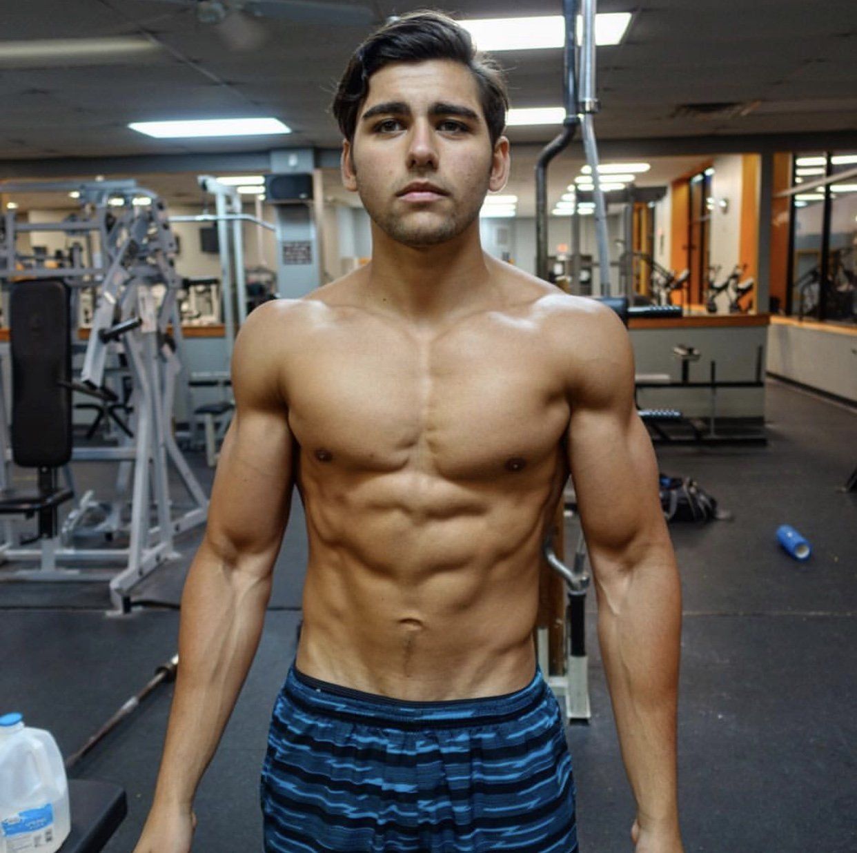 A shirtless man is standing in a gym holding a barbell