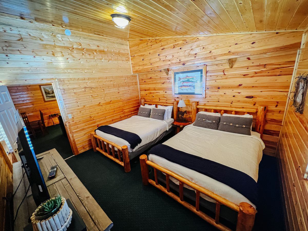 Cozy cabin bedroom with two beds, wood paneling, TV, and a view into another room.