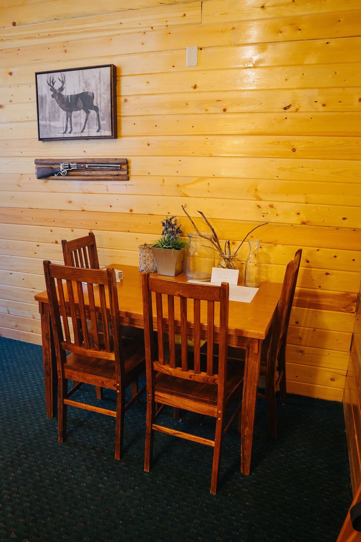 Wooden dining table and chairs in front of a wood-paneled wall, decorated with a deer picture and a shelf.