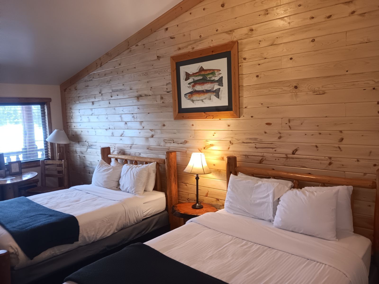 Two beds in a wood-paneled room, framed fish artwork, bedside lamps, window with blinds.