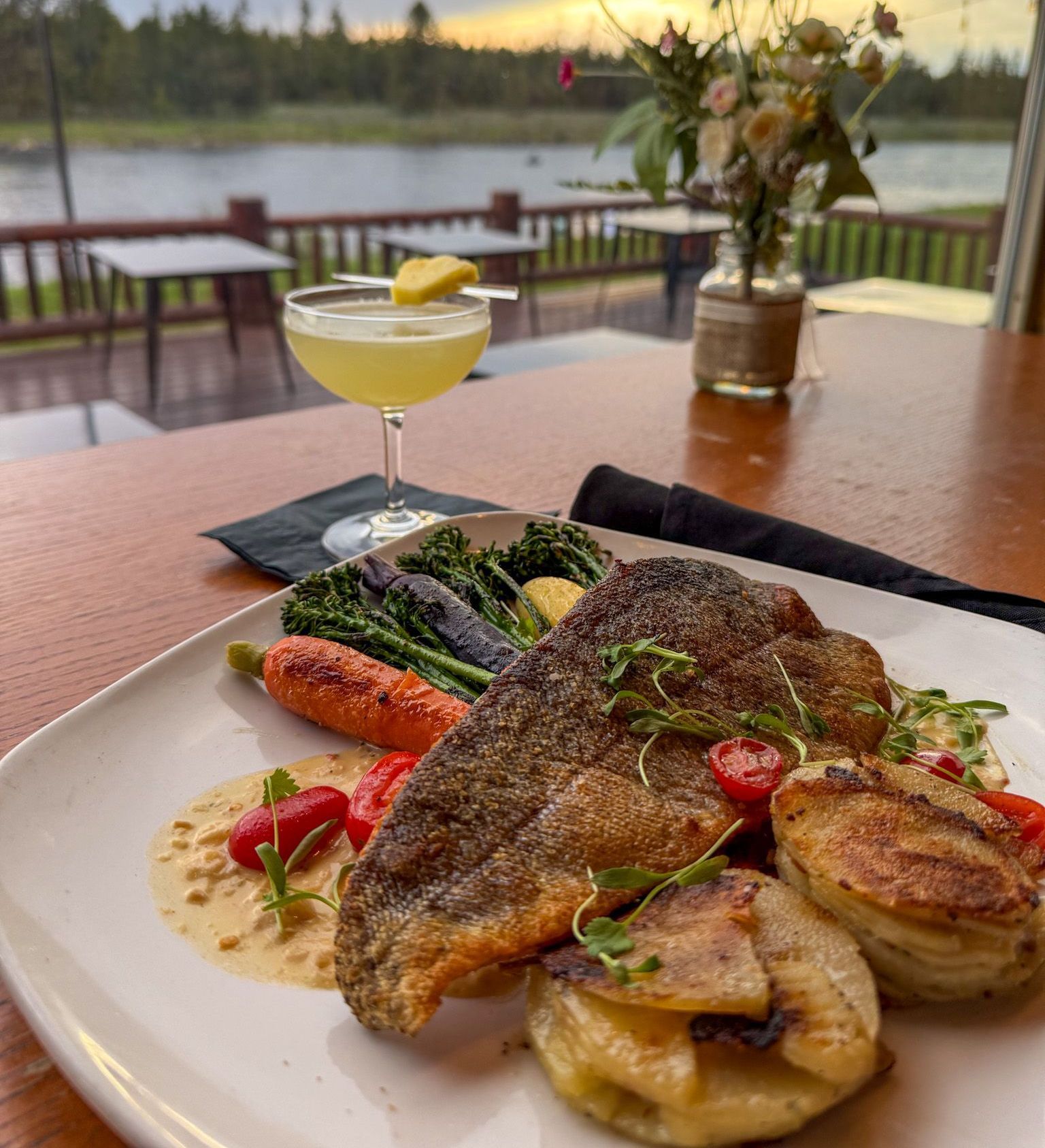 Plate of seared fish with vegetables and cocktail on a restaurant table overlooking a lake at sunset.