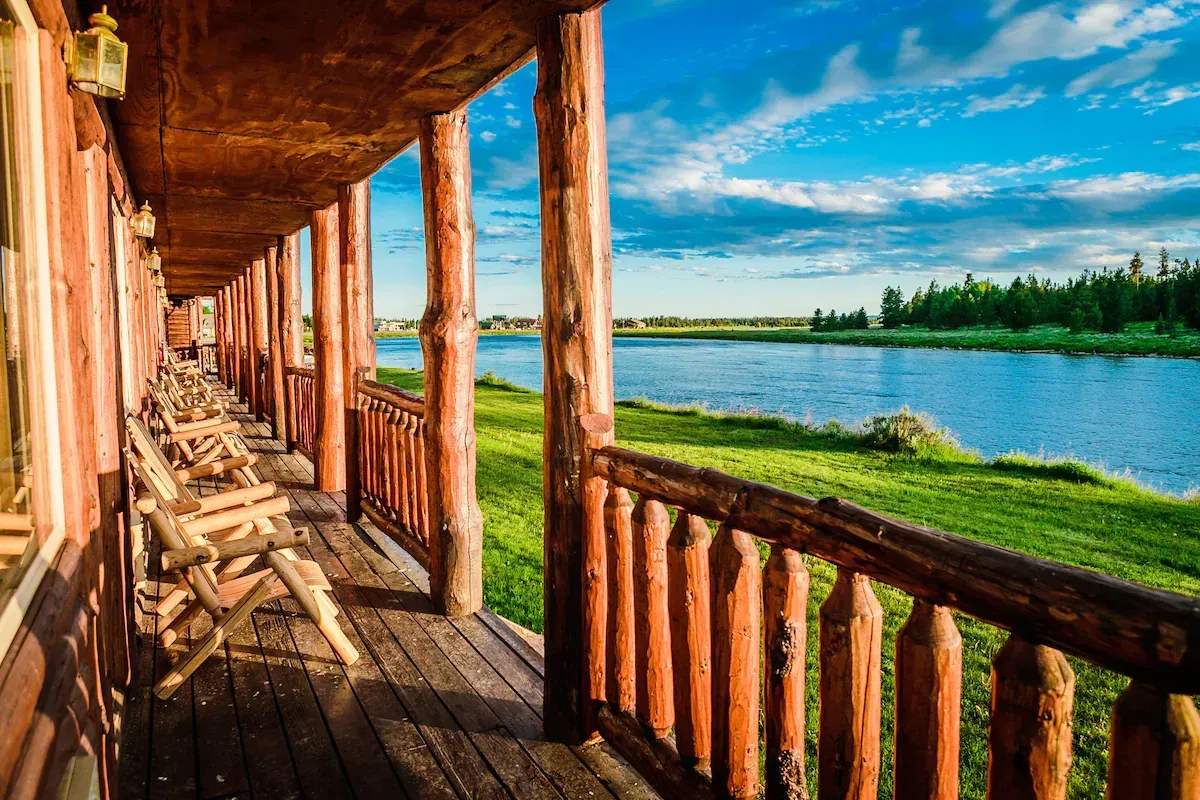 Wooden porch overlooking a river, sunny day. Rustic lodge with chairs. Green grass, blue sky.