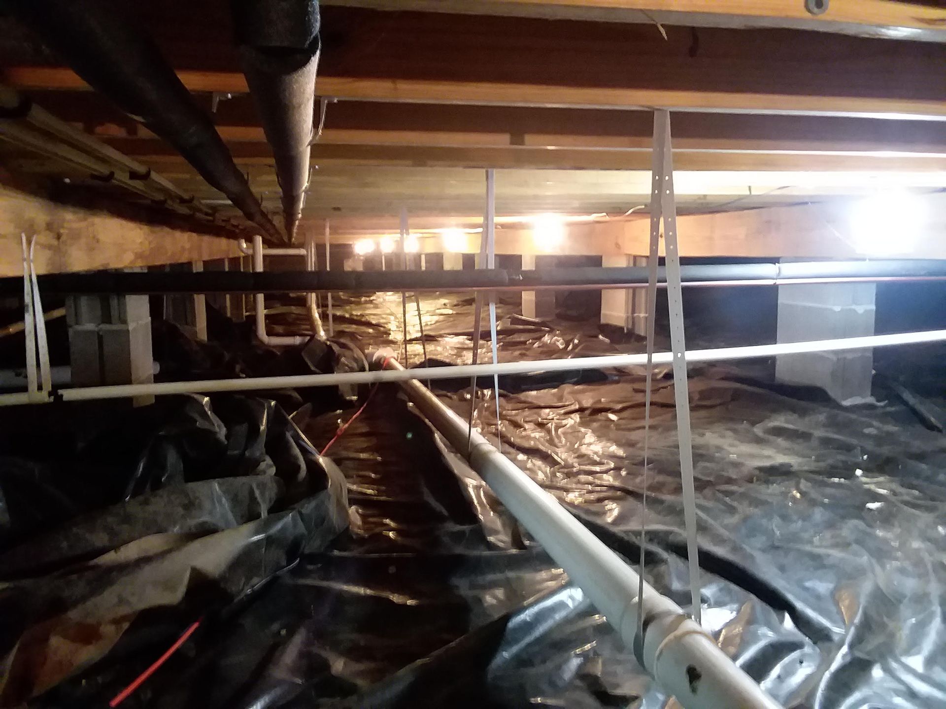 Crawlspace with exposed pipes and plastic vapor barrier under a wooden floor, lit by a flashlight