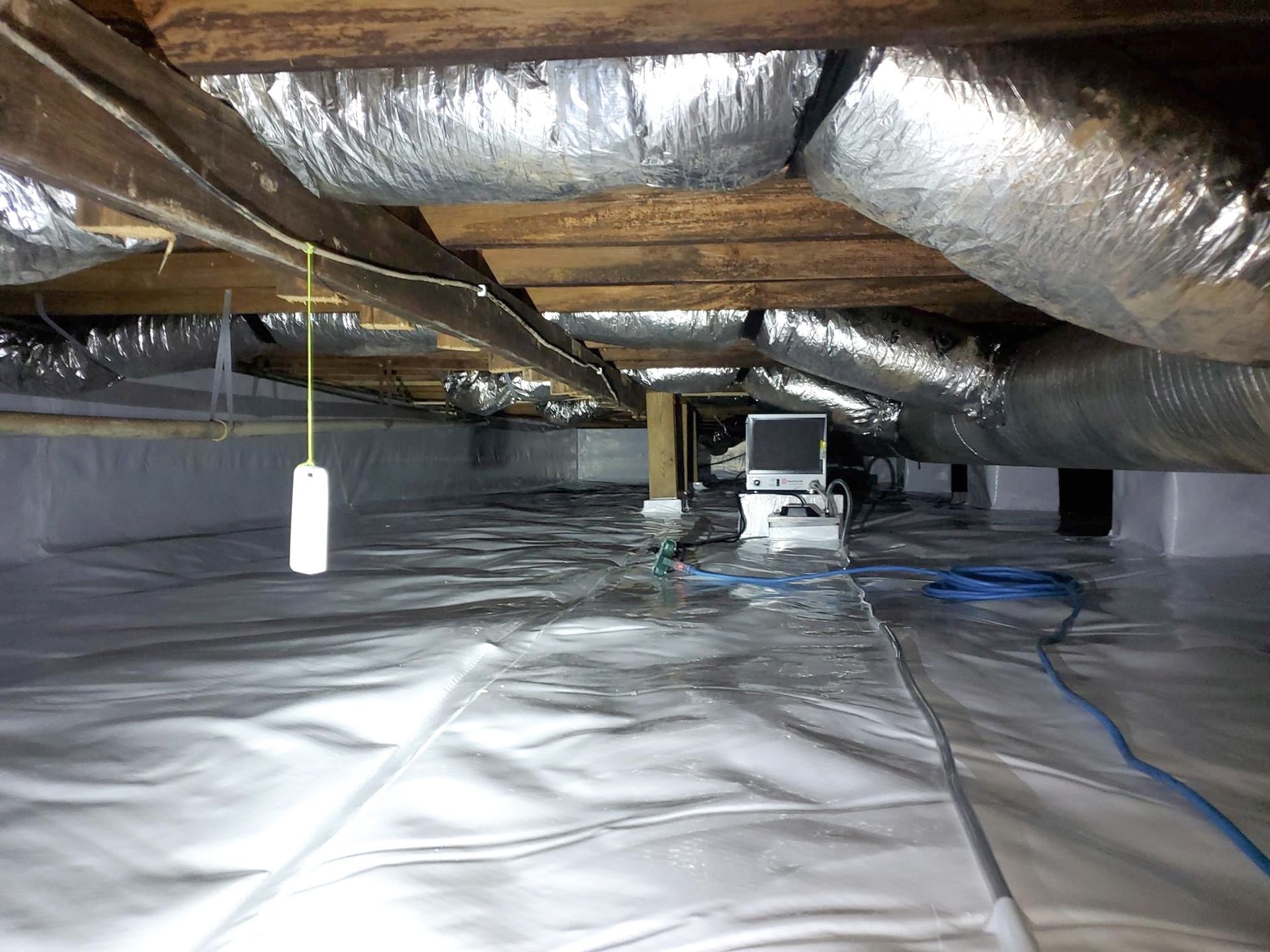 Crawlspace with insulated ducts, exposed wooden joists, plastic floor sheeting, and a portable work light