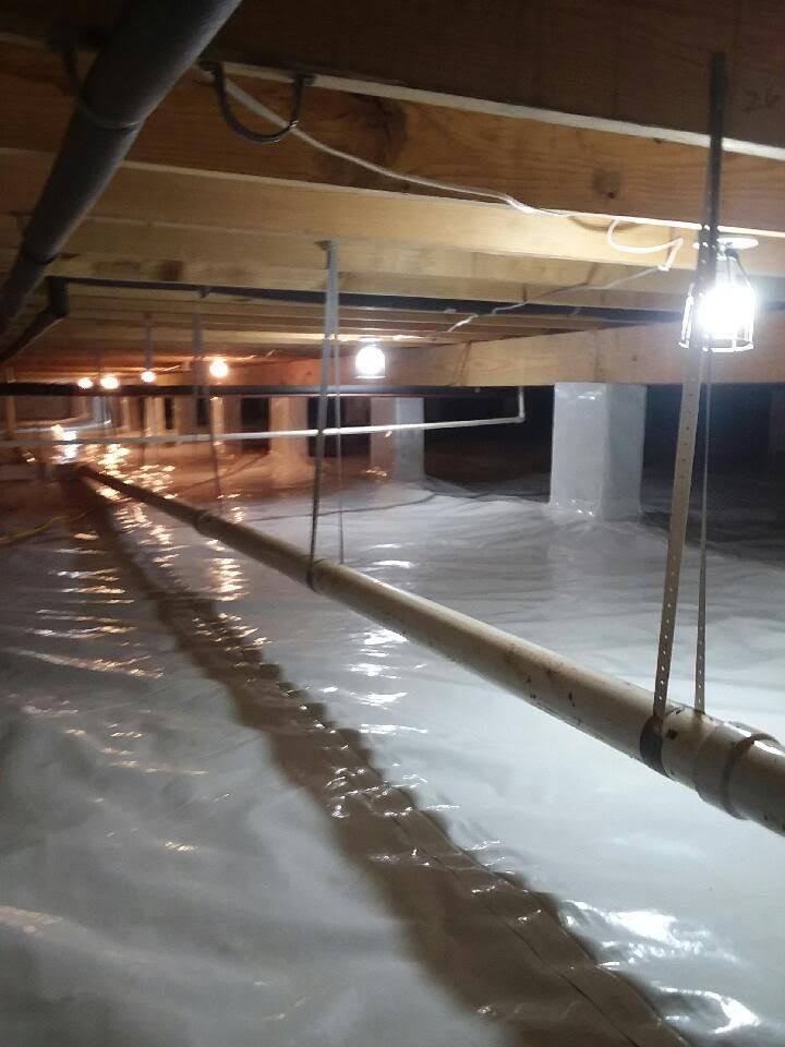Indoor crawlspace with reflective vapor barrier, wooden beams, pipes, and bright hanging work lights