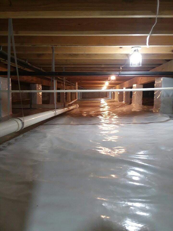 Crawlspace under a house with white plastic vapor barrier, exposed beams, pipes, and a hanging light