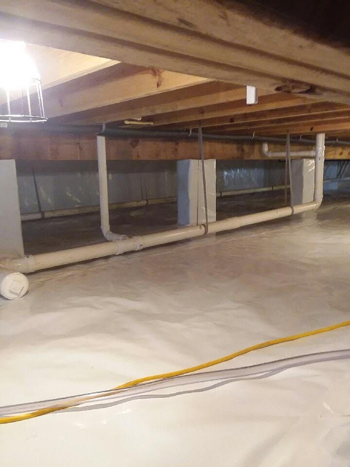 Dim crawl space under a house with wooden beams, white support posts, and a yellow cable on the floor.