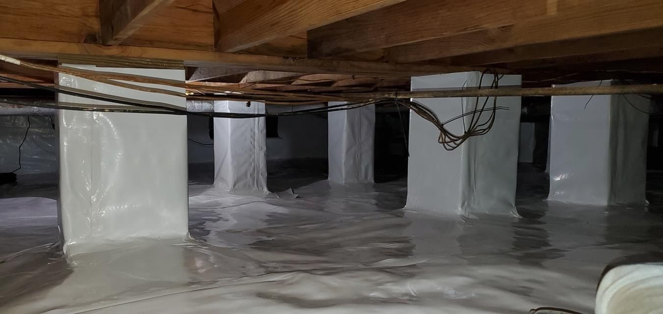 Plastic-covered crawl space under wooden beams with support posts and pipes