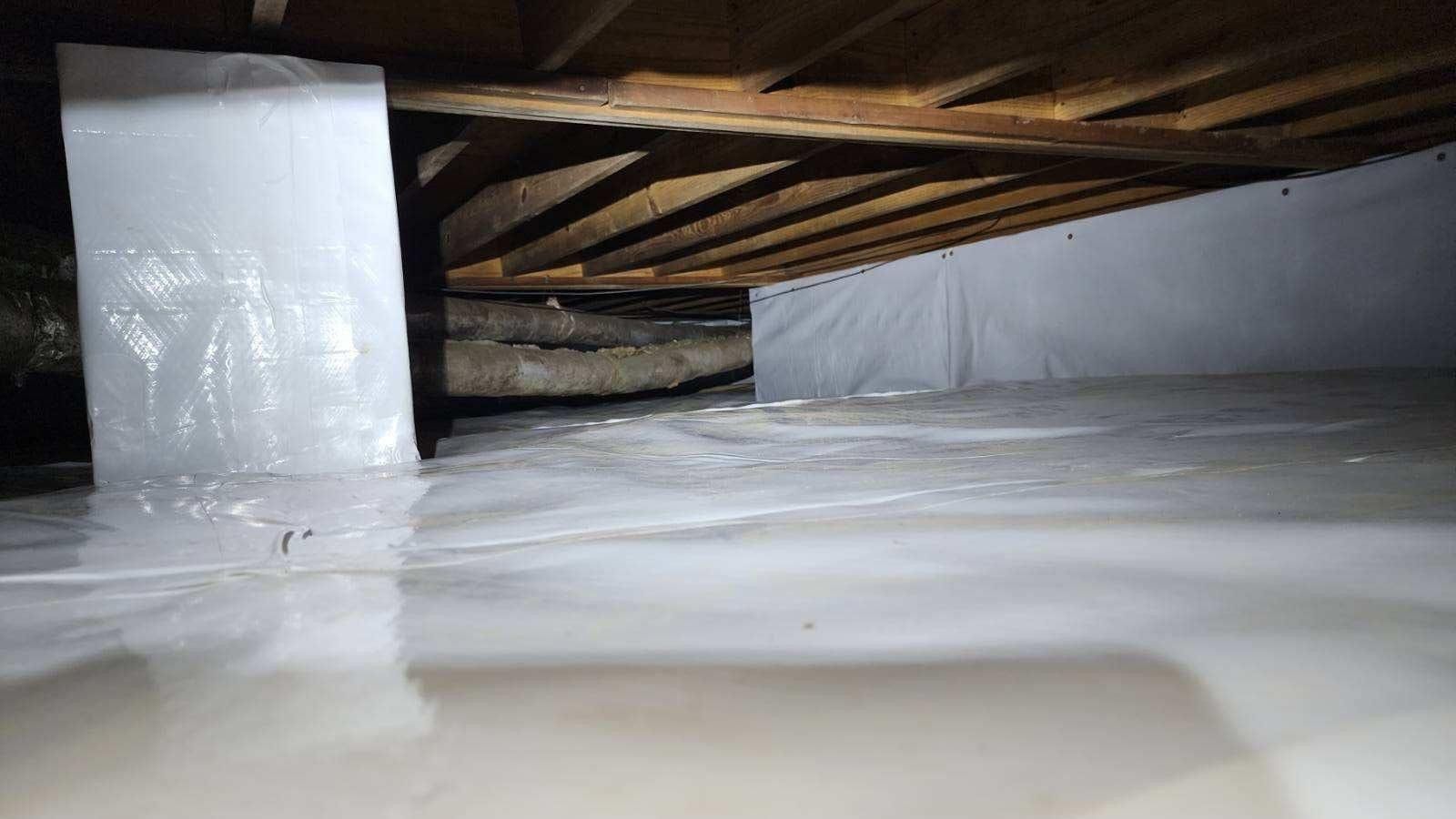 Inside a crawlspace, white vapor or insulation beneath wooden floor joists and support posts