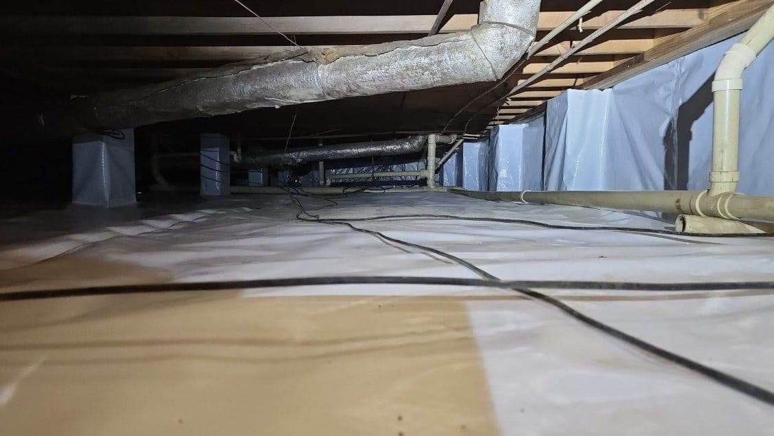 Crawlspace under a house with exposed pipes, insulation, and ductwork along the floor and ceiling.