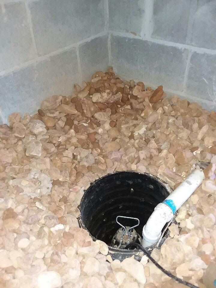 Black drainage pipe surrounded by tan rocks in a concrete corner, with a white PVC elbow attached.