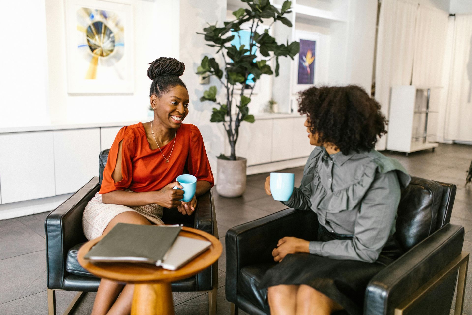 Two women are sitting in chairs talking to each other while drinking coffee.