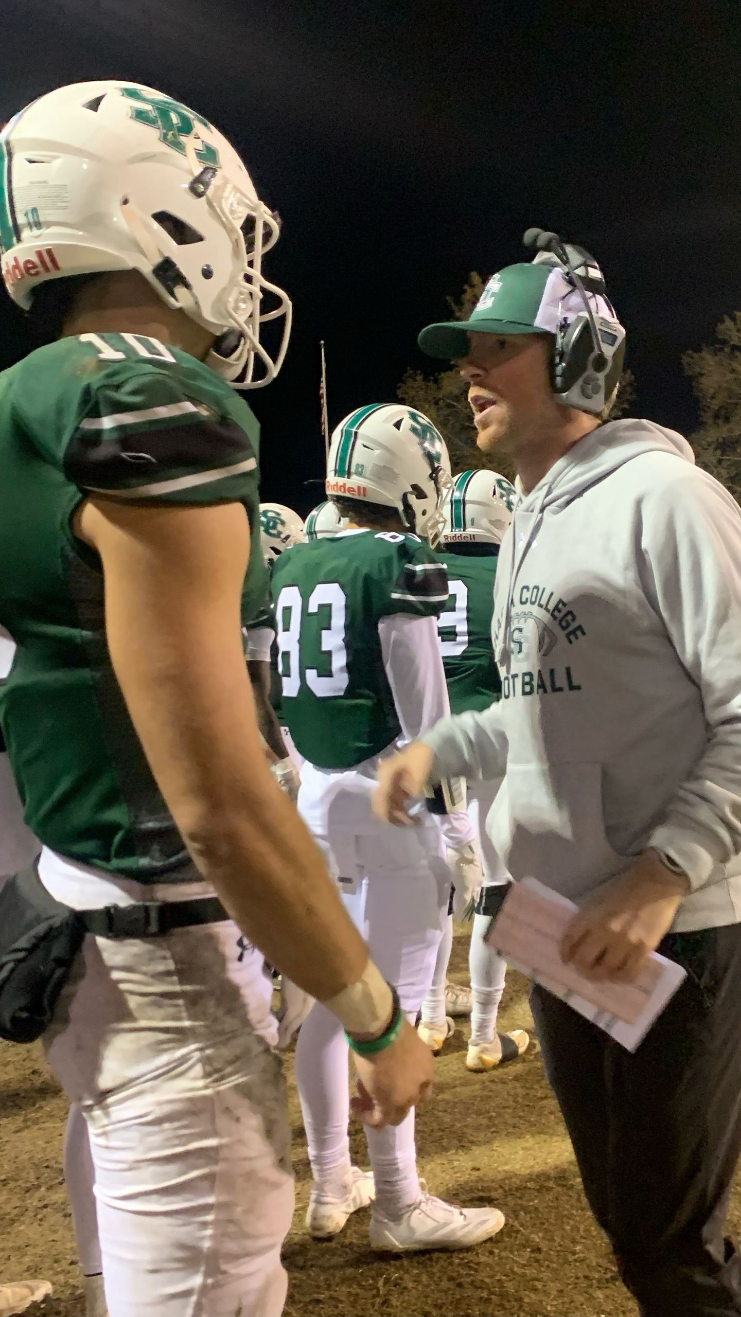 Football coach talking with a player on a night field. The coach wears a green cap and sweatshirt, the player in a green and white uniform.