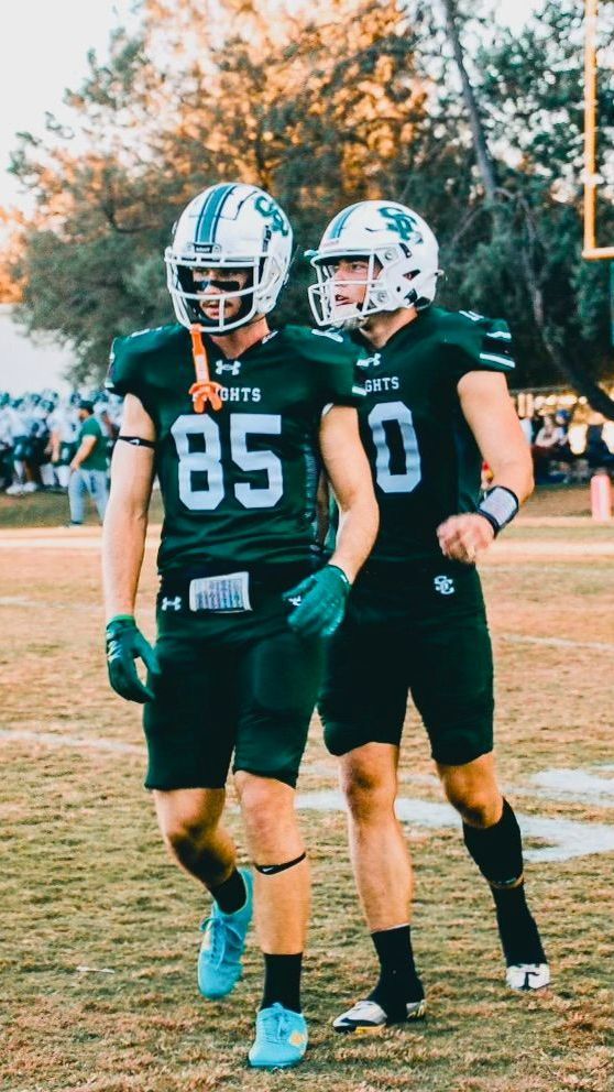 Two football players in green uniforms walk on a field; one with the number 85, the other with 10.