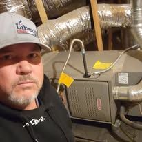 A man wearing a hat is standing next to a furnace in a basement.