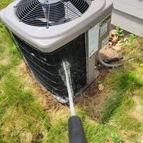 A person is cleaning an air conditioner with a brush in the grass.