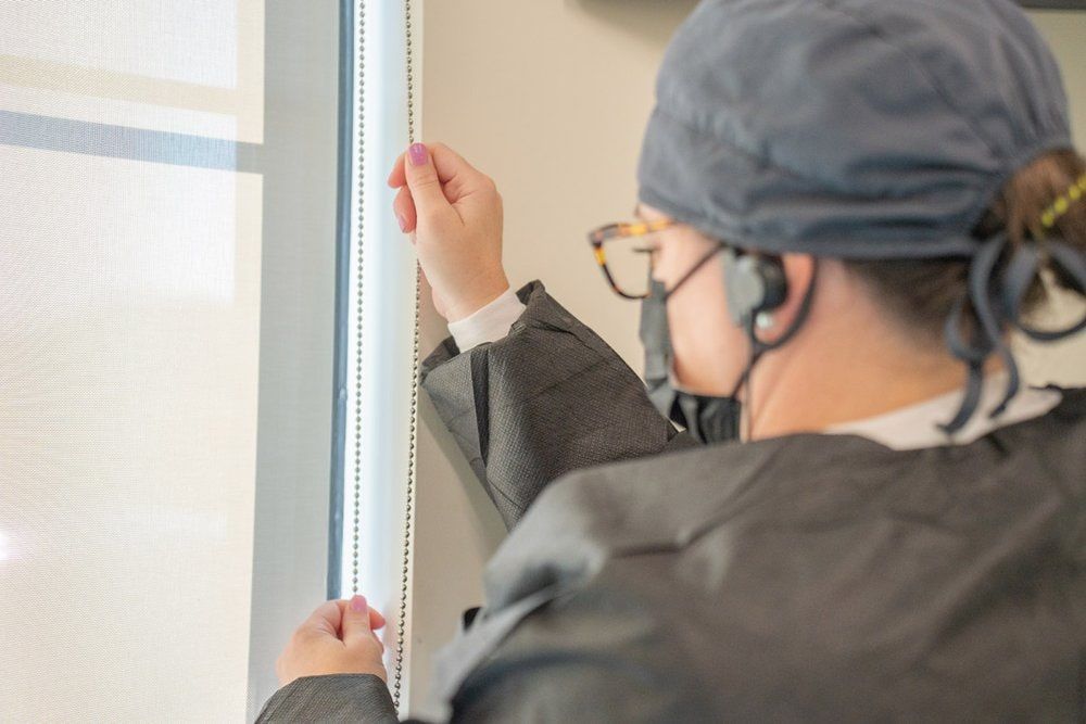 A woman wearing a mask and gown is adjusting a solar shade on a window.