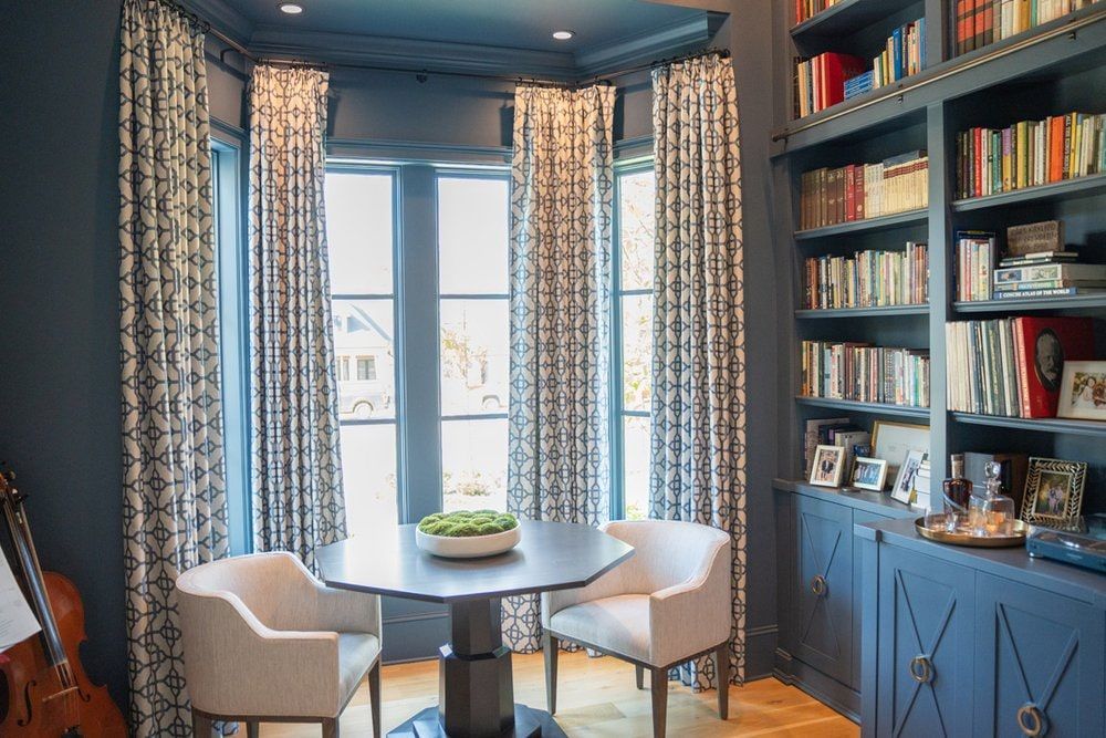 There is a table and chairs in the middle of the room with custom drapes over the windows.