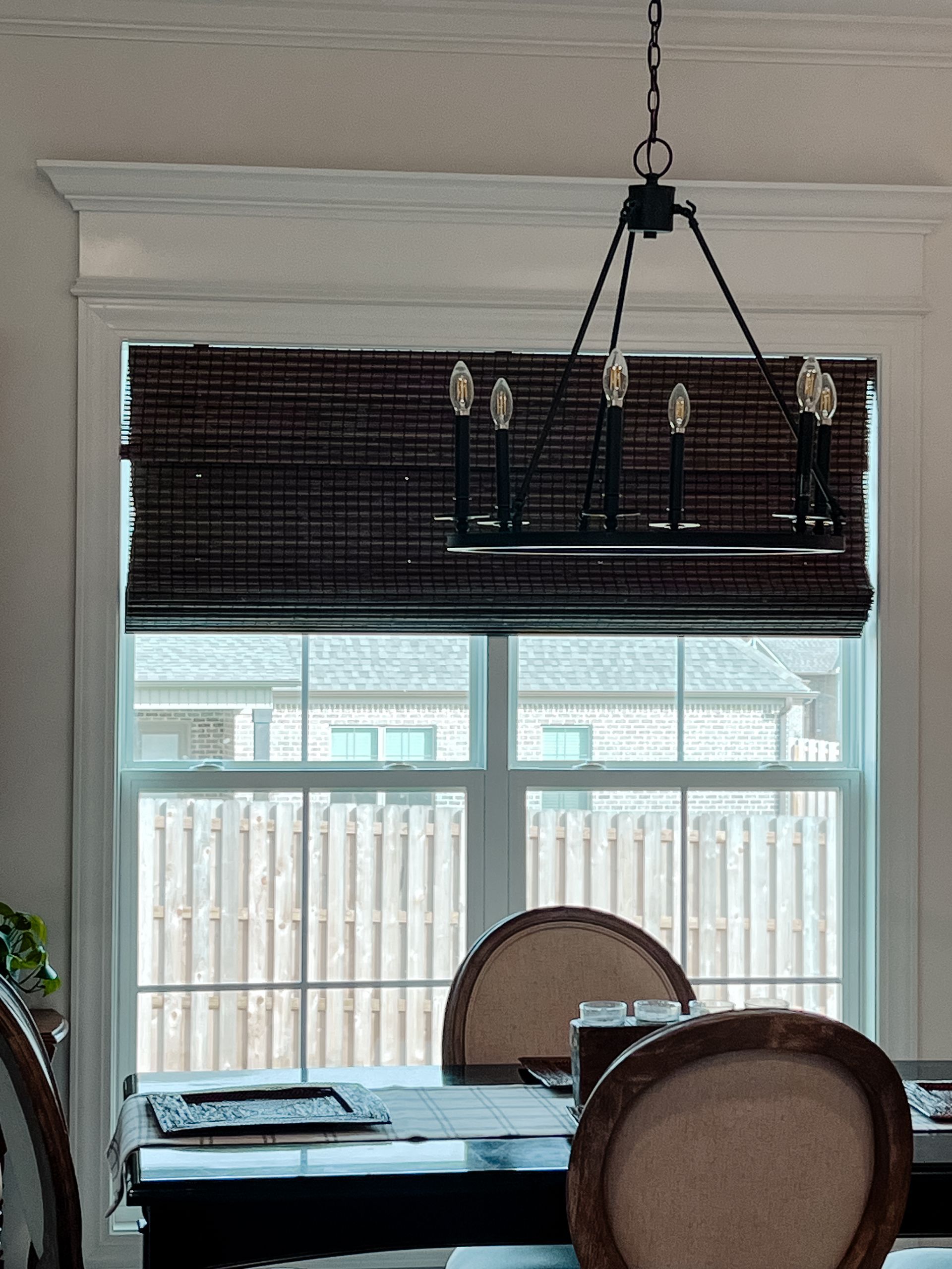 A dining room with a table and chairs and a chandelier hanging from the ceiling and window with natural women shade.