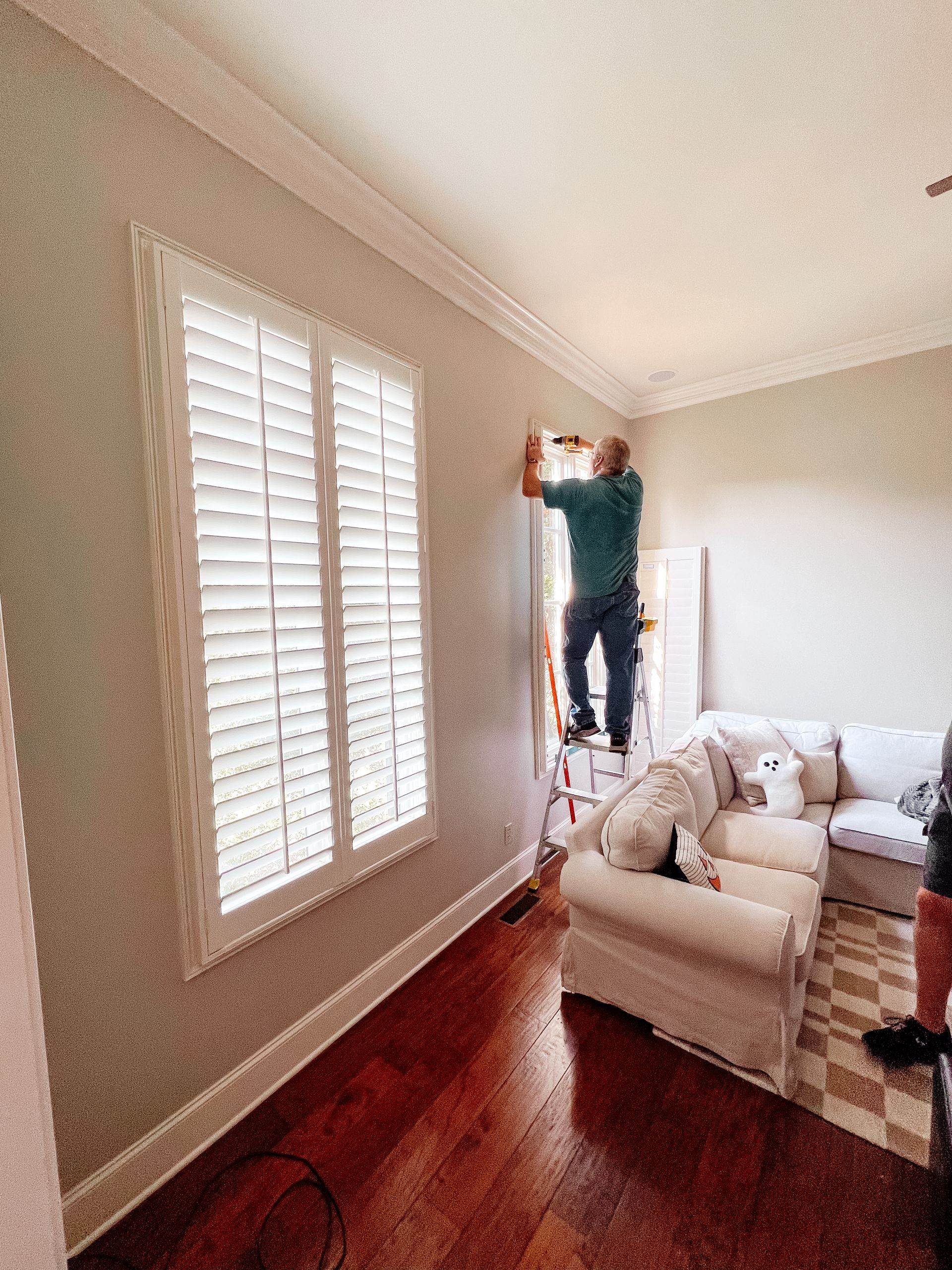 A man is standing on a ladder installing a wooden shutter on a window in a living room.