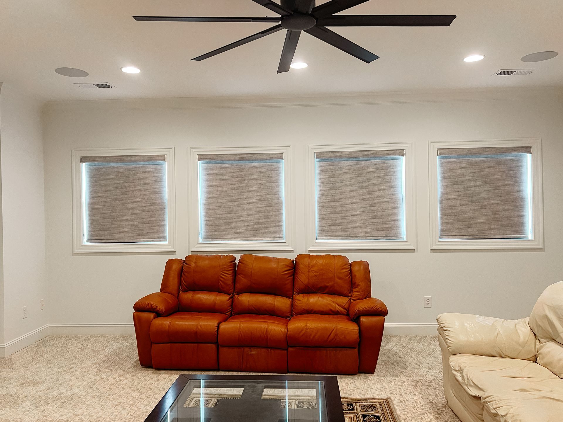 A living room with a brown couch and a ceiling fan and woven shade on the windows.