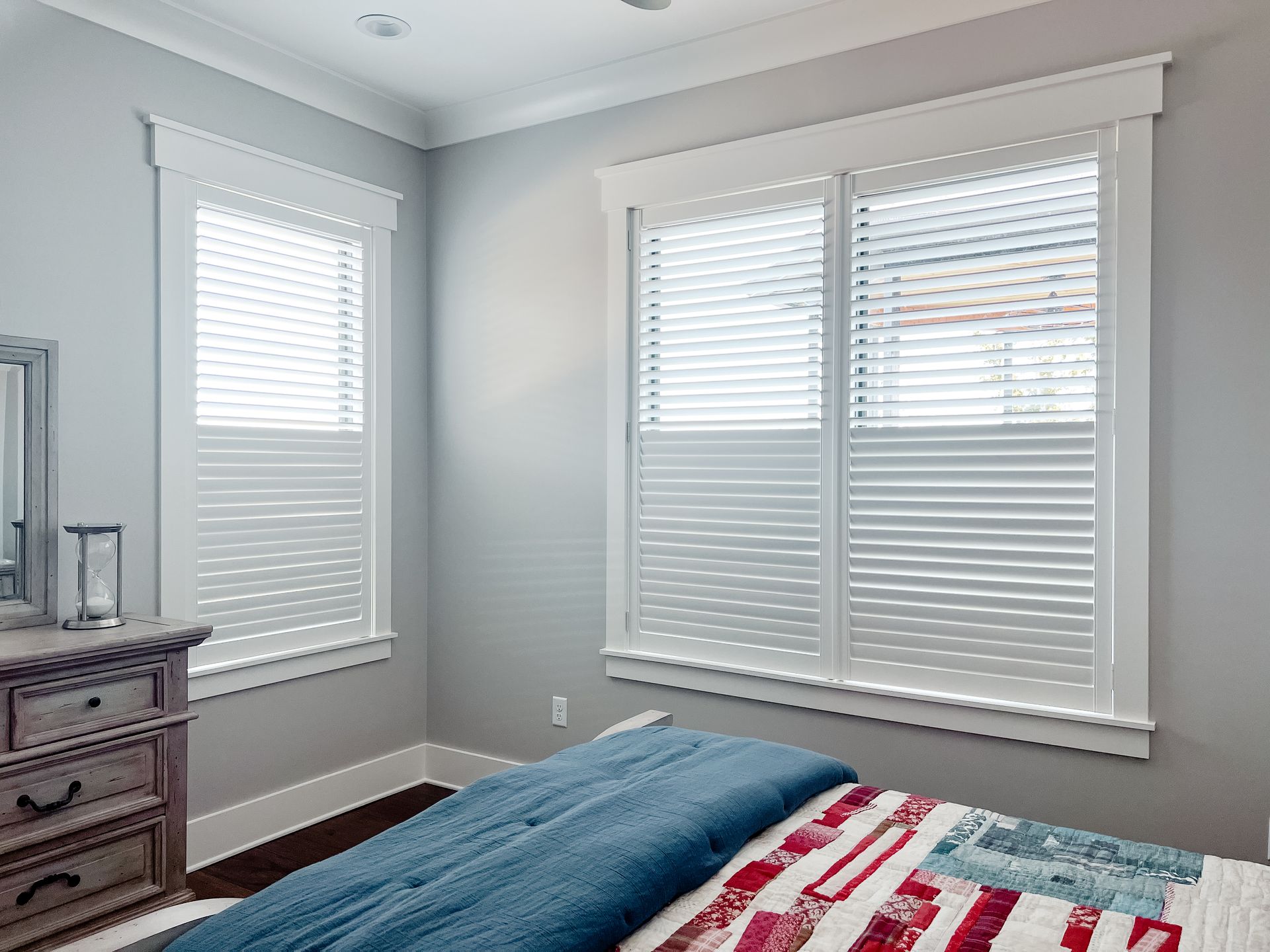 A bedroom with a bed , dresser , mirror and two windows with wooden shutters.