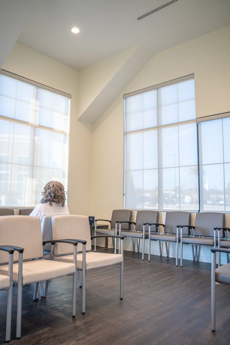 A woman is sitting in a waiting room with chairs and windows with solar shades.