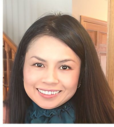 Woman with dark hair smiling at the camera; she's wearing a teal top in a home setting.