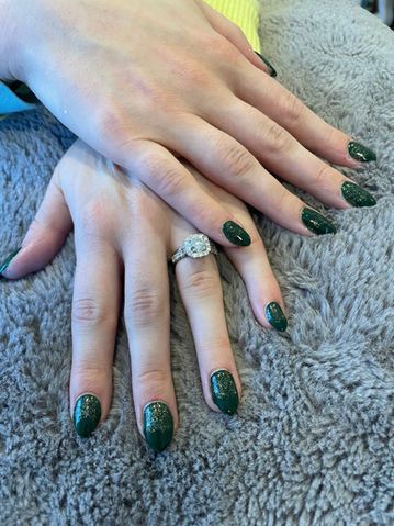 Hands with dark green polished nails, one wearing a diamond engagement ring, resting on gray fuzzy fabric.