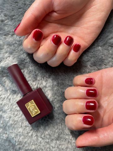 Two hands with short, dark red manicured nails next to a bottle of matching nail polish on a gray surface.
