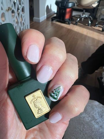 Hand holding a green nail polish bottle, light pink nails, one decorated with a Christmas tree design.