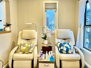 Two cream-colored pedicure chairs in a nail salon with small table of products between them; window visible.