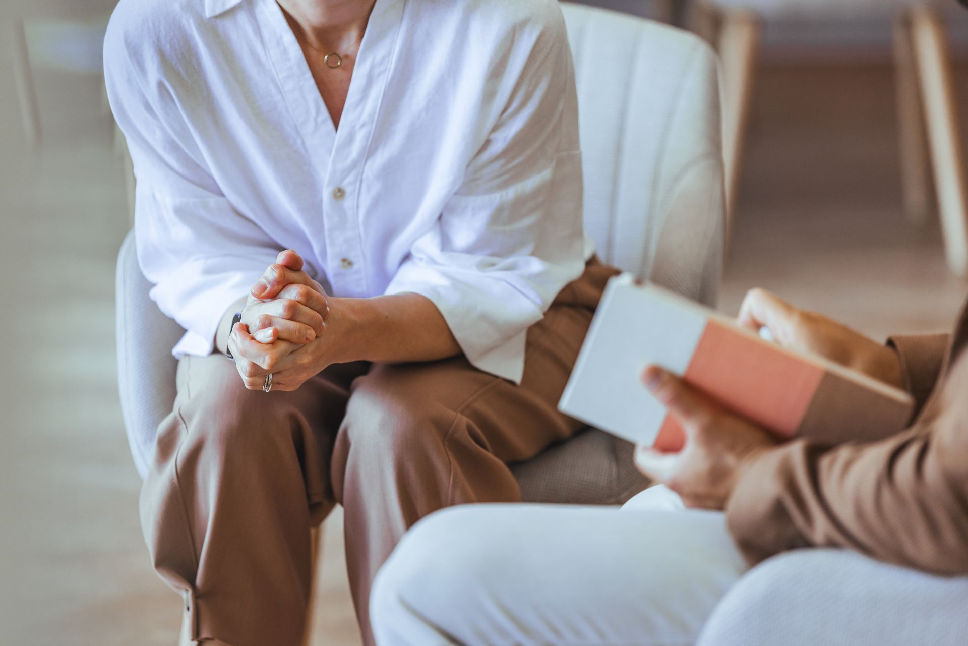 Person in white shirt and brown pants, hands clasped, facing another person holding a book, seated in a chair.