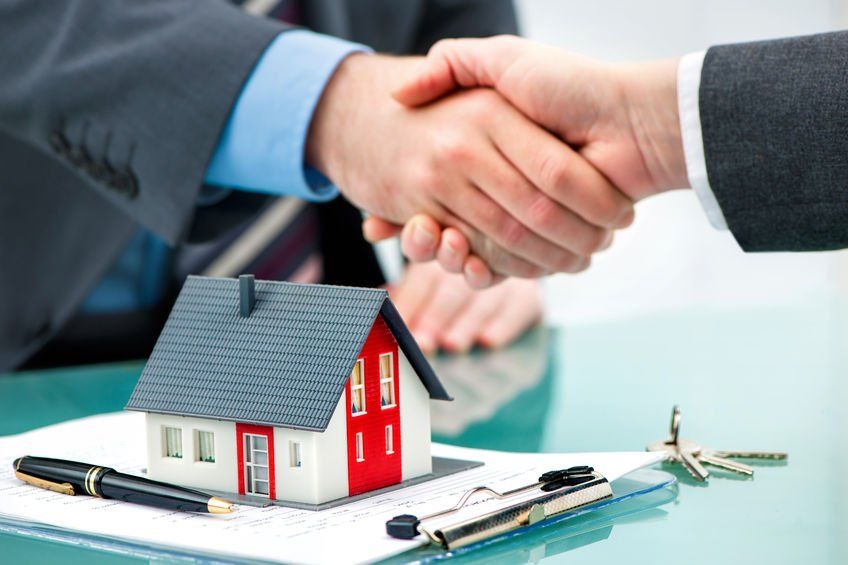 Two people are shaking hands over a model house on a clipboard.