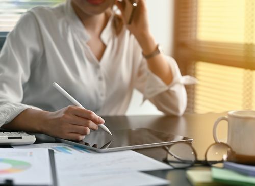 A woman is sitting at a desk using a tablet and talking on a cell phone.